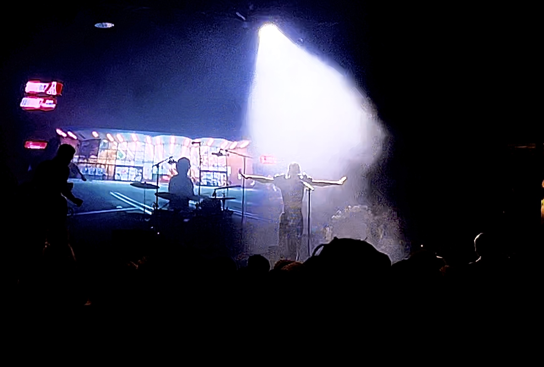The lead singer under a spotlight, obscured by fog and backlit so she is mostly only visible through her silhouette. The drummer is silhouetted next to her.